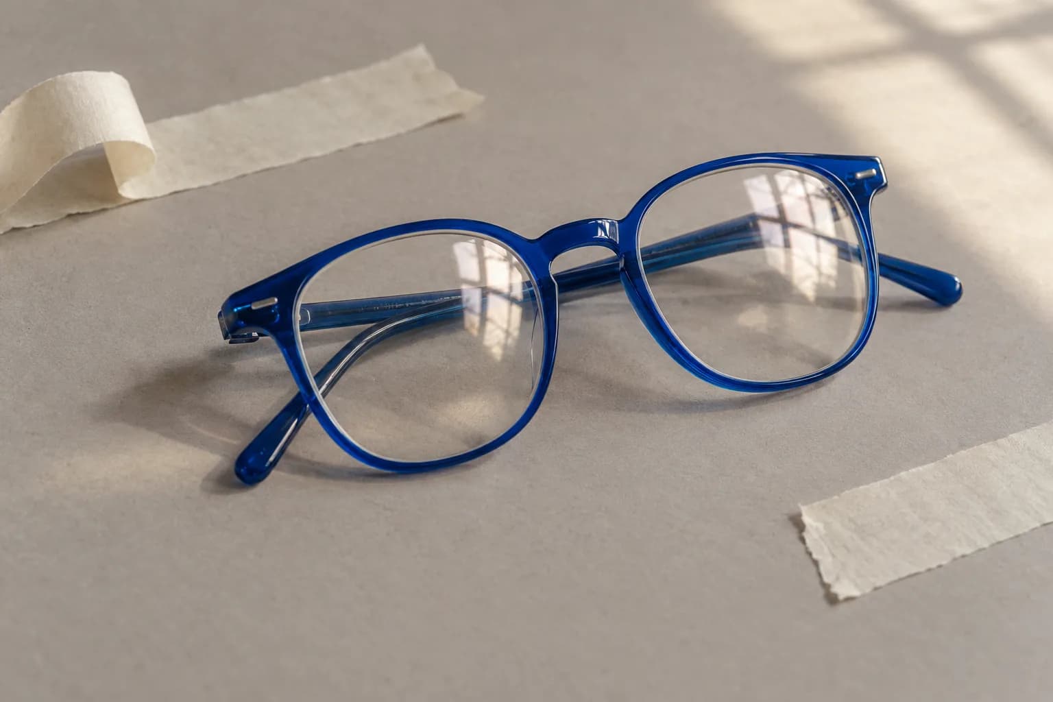 Close-up of a pair of eyeglasses with electric-blue frames on warm grey paper, window reflections in the lenses, strips of off-white tape nearby.