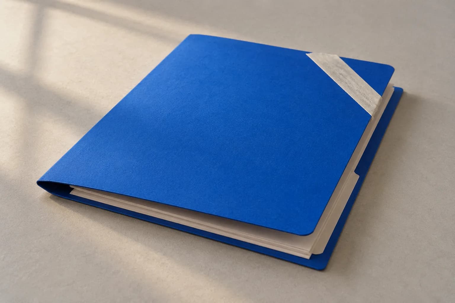 An electric-blue cardboard document folder, slightly open, with blank white pages inside and a strip of off-white tape across the corner, on warm grey paper.