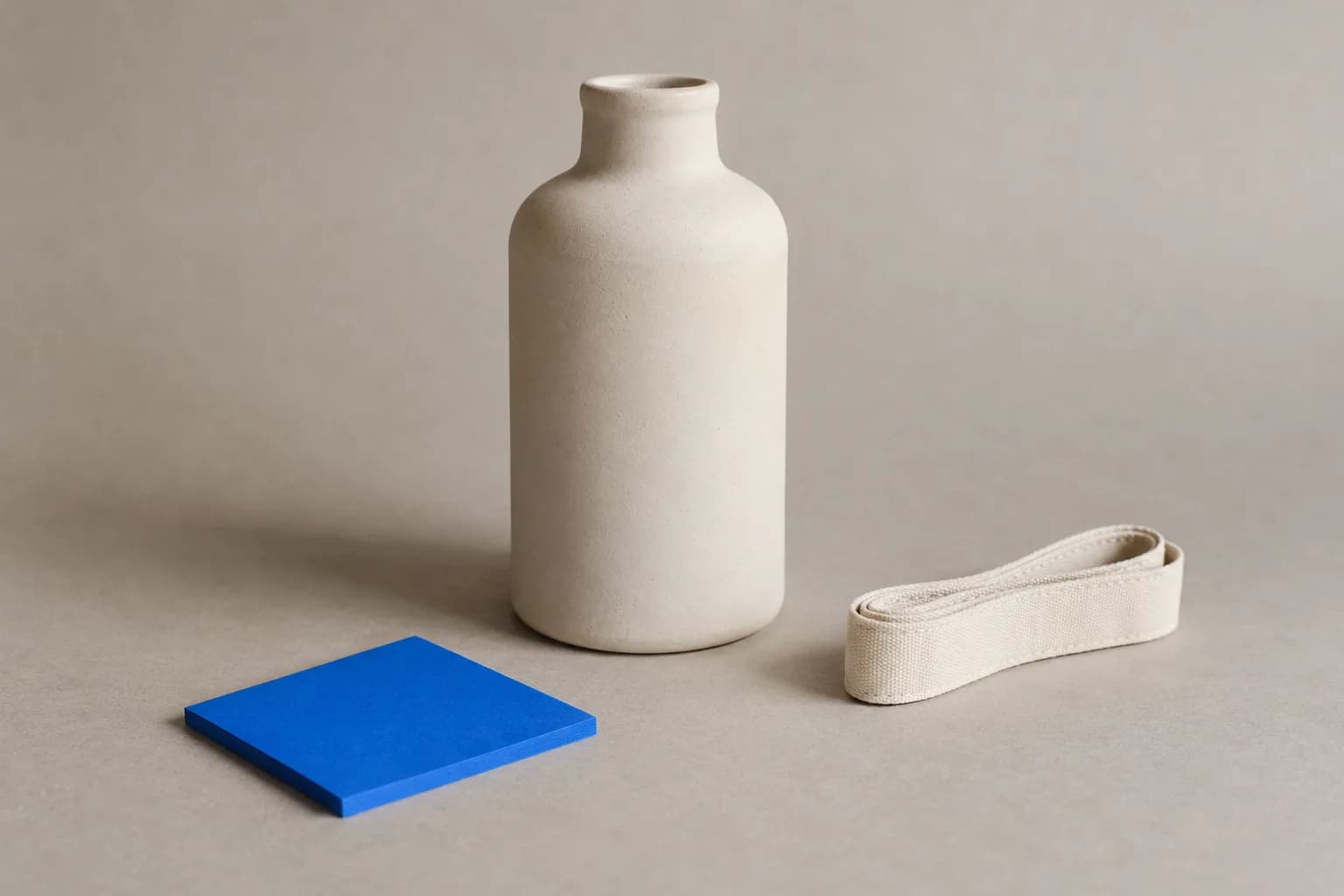 A plain matte ceramic bottle standing on warm grey paper beside a folded cloth tape measure and one electric-blue square sticky note.
