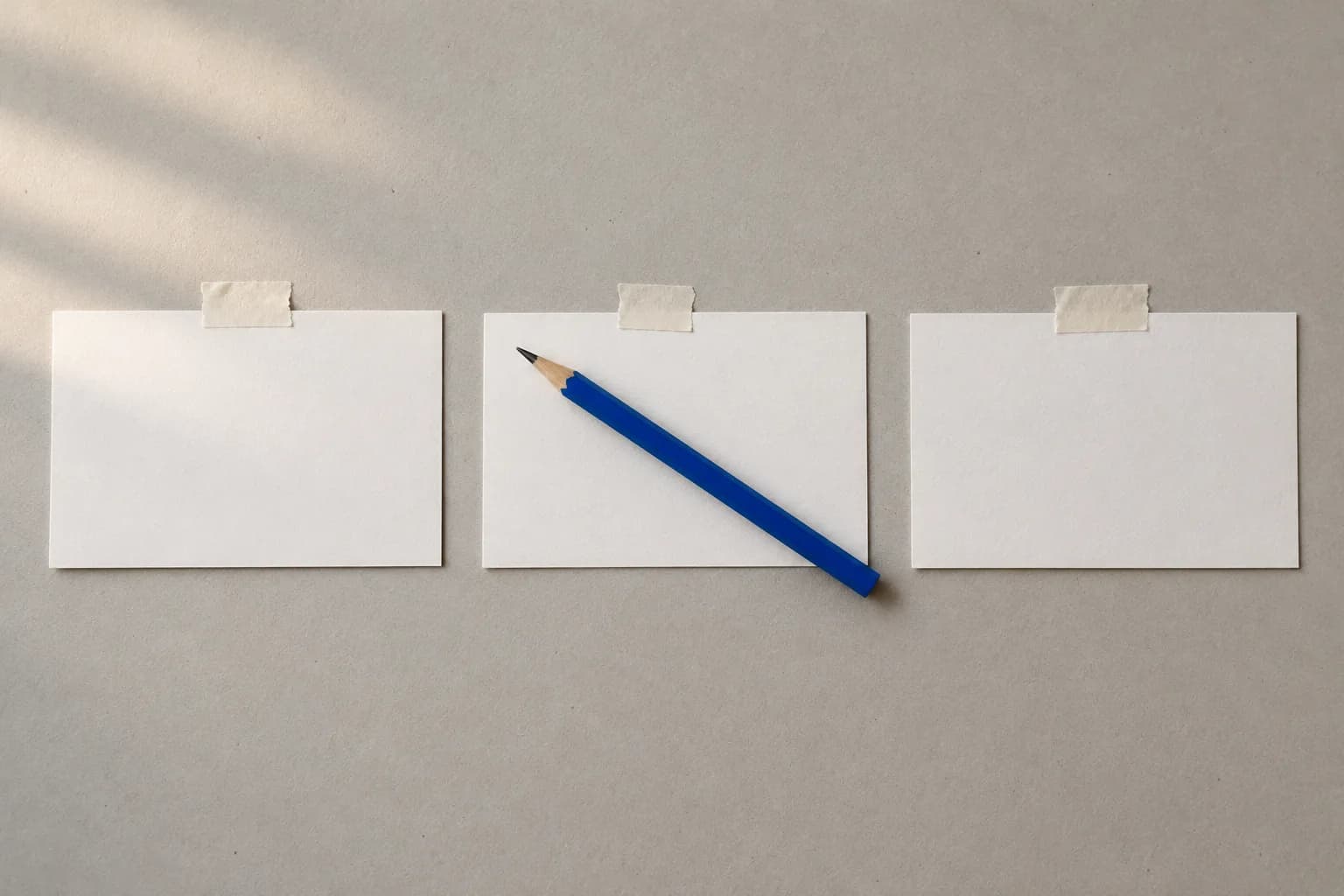 Three blank white index cards laid in a row on warm grey paper with a single electric-blue pencil resting across the middle one.